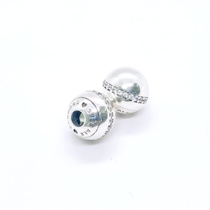 May include: Two spherical silver charms featuring a central band of clear pavé stones. The side of one charm is engraved with the text PANDORA, a heart symbol, 5925, and ALE, indicating sterling silver composition and brand authenticity.