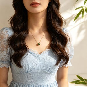 Puede incluir: Un collar dorado con un colgante redondo verde oscuro. El colgante está engastado en un marco dorado. El collar se lleva sobre un vestido de encaje azul claro. La imagen está bien iluminada, destacando la joya y los detalles del vestido.