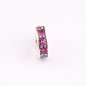 Puede incluir: Un abalorio de plata con una hilera de piedras preciosas cuadradas de color rosa magenta vibrante engastadas en una banda metálica. El diseño incluye pequeñas cuentas de plata que separan cada piedra cuadrada, creando un aspecto pulido.