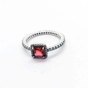 May include: A silver-toned ring featuring a square-cut red gemstone at the center, surrounded by a halo of small clear stones. The band is also encrusted with a series of small clear stones extending along the sides of the metal ring structure.