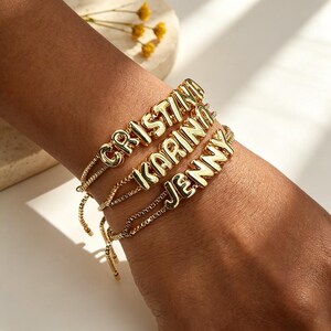 Op de afbeelding: Goudkleurige armbanden met de namen "CRISTINA", "KARINA" en "JENNY" in blokletters. De armbanden zitten om een pols, met een fijne ketting en een verstelbare sluiting. De achtergrond is een zachte, neutrale kleur.