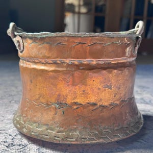 May include: A copper-colored, antique-style bucket with two small handles. The bucket has a flared base and is adorned with decorative stitching around the top and bottom. The metal has a weathered appearance, suggesting age and use.