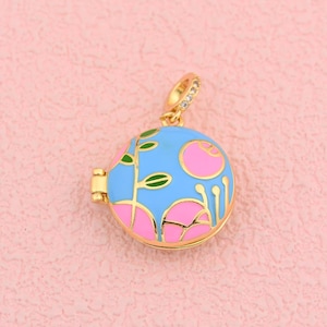 May include: A round, gold-toned pendant with a blue enamel background and pink floral design. The pendant has a hinged opening and a gold bail with small, clear stones. The floral design includes pink flowers and green leaves.