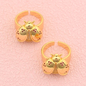 May include: Two gold-colored adjustable rings, each shaped like a ladybug. The ladybug's wings have red accents. The rings are on a pink background. The rings are likely made of metal.