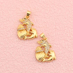 May include: Two gold-colored dolphin pendants with clear crystal accents. Each pendant features a dolphin leaping over a seashell. The pendants have a loop at the top for attaching to a chain. The background is a light pink color.
