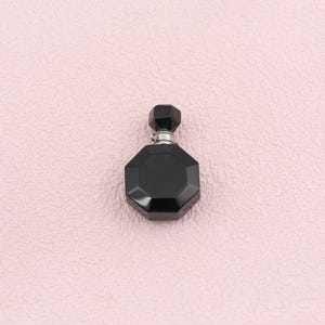 Puede incluir: Un pequeño frasco de perfume negro de forma octogonal con una superficie facetada y un tapón negro a juego de forma geométrica hexagonal. El frasco presenta un cuello de metal plateado que conecta el cuerpo principal con el tapón.