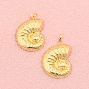 May include: Two gold-colored pendant necklaces in the shape of a nautilus shell. The pendants have a textured surface and a loop at the top for a chain. The background is a solid light pink color.
