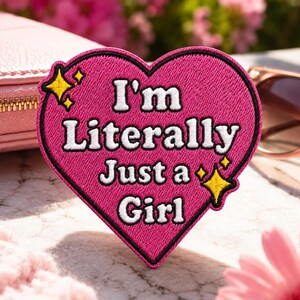 May include: A heart-shaped embroidered patch in hot pink with a black border. The patch features the white text "I'm Literally Just a Girl" in a bold font, accented by two yellow four-pointed star icons on the left and right sides.