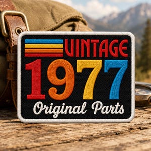 May include: A rectangular embroidered patch with a black background featuring the red text VINTAGE above the large colorful numbers 1977 in red, orange, yellow, and blue. The white text Original Parts is below, with horizontal stripes above.