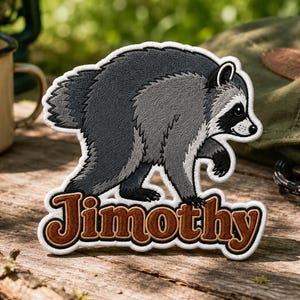May include: An embroidered patch featuring a gray and black raccoon walking toward the right. Below the raccoon, the name Jimothy is stitched in brown letters with a white outline. The patch has a white border and a die-cut shape.