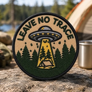 May include: A circular embroidered patch with a tan background and a black border. It features a gray UFO hovering above a dark green forest, emitting a yellow light beam that illuminates a small tent. The text LEAVE NO TRACE is stitched in black.