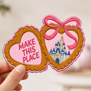 May include: An embroidered patch shaped like two connected hearts with scalloped gold borders and pink edges. The left heart features the pink text "MAKE THIS PLACE" and the right heart displays a castle illustration topped with a pink bow.