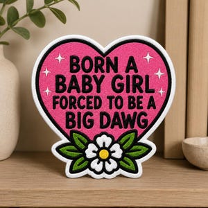May include: A heart-shaped embroidered patch with a pink background and black text that reads BORN A BABY GIRL FORCED TO BE A BIG DAWG. The design features white four-pointed stars and a white flower with a yellow center and green leaves at the base.