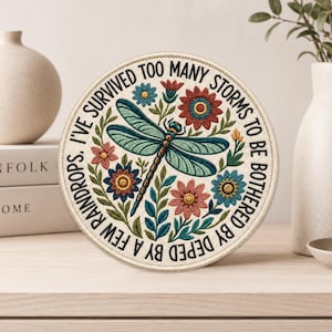 May include: A circular embroidered patch featuring a central teal dragonfly surrounded by colorful red, pink, and blue flowers and green leaves. The text "I'VE SURVIVED TOO MANY STORMS TO BE BOTHERED BY A FEW RAINDROPS" is stitched in black around the border.