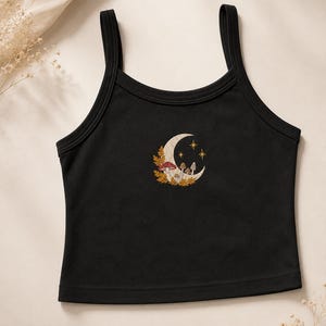 May include: A black sleeveless crop top featuring a central graphic print of a crescent moon, small mushrooms, and golden leaves. The moon design includes a red-capped mushroom and two small stars, all centered on the chest of the garment.