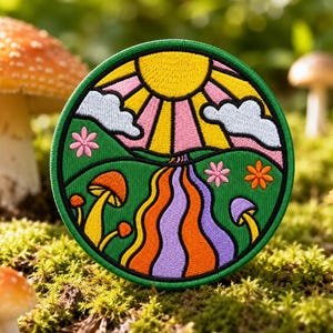 May include: A circular embroidered patch featuring a vibrant landscape design. The scene depicts a yellow sun with pink rays, light blue clouds, green hills, and a winding orange and purple path. Small mushrooms and colorful flowers are stitched throughout.