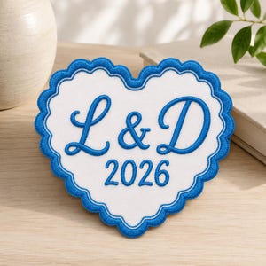 May include: A heart-shaped patch with a white center and a scalloped blue border. The embroidery features the blue letters L & D in a cursive script, positioned above the blue embroidered year 2026, centered on the white fabric background.