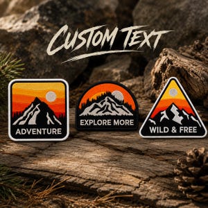 May include: Three embroidered patches featuring mountain landscapes with orange and yellow sunset skies. The square patch says ADVENTURE, the semi-circular patch says EXPLORE MORE, and the triangular patch says WILD & FREE in white text.