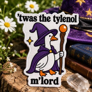 May include: A white embroidered patch featuring a goose wearing a purple wizard hat and cape, holding a wooden staff with an orange orb. The text 'twas the tylenol' is above the goose, and 'm'lord' is below, all in black, bold, sans-serif font.