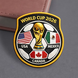 May include: Embroidered patch featuring the World Cup 2026 logo. The design includes a soccer ball with the flags of the USA, Mexico, and Canada, along with the World Cup trophy. The patch has a black background with gold trim.