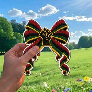 May include: A decorative bow-shaped patch featuring a black power fist in the center. The bow has red, black, and green stripes with gold trim. The patch is held in front of a sunny, green field with a blue sky.