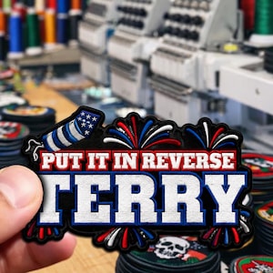 May include: Embroidered patch with the text "PUT IT IN REVERSE TERRY" in white, red, and blue. The patch features an American flag design and fireworks. The patch is black with a white border.