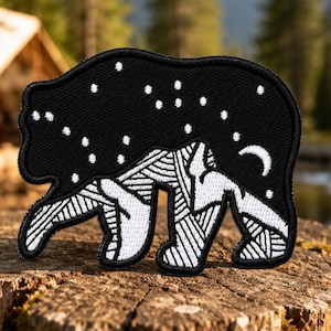 May include: A black and white embroidered patch shaped like a bear silhouette. The design features a starry night sky above a mountain range depicted with white diagonal lines. The bear's body is filled with these mountain patterns and stars.
