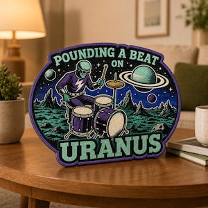 May include: An embroidered patch featuring a green alien playing a drum set on a rocky planet surface. The design includes a Saturn-like planet, stars, and the text POUNDING A BEAT ON URANUS in light green letters against a dark blue background.