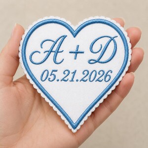 May include: A heart-shaped white fabric patch with a scalloped edge and blue embroidered border. The center features the blue embroidered letters A + D in a cursive script, with the date 05.21.2026 embroidered in blue below the letters.