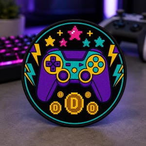 May include: A circular embroidered patch featuring a purple and teal video game controller at the center. The design includes yellow lightning bolts, pixelated stars in pink and teal, and three gold pixelated coins with the letter D in the center.