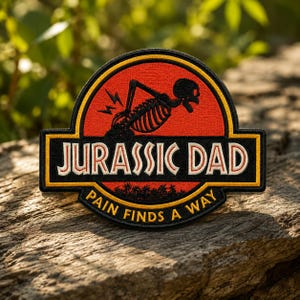 May include: Embroidered patch with the text "JURASSIC DAD" in white letters on a black and gold rectangular background. The patch features a red circle with a skeleton and the words "PAIN FINDS A WAY".