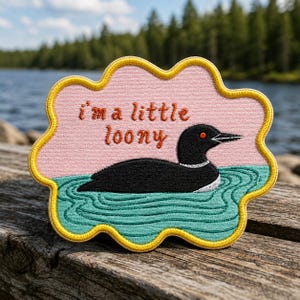 May include: A cloud-shaped embroidered patch with a yellow border, featuring a black loon swimming in teal water against a light pink background. The text "i'm a little loony" is stitched in orange letters above the bird, which has a red eye.