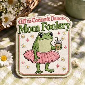May include: A rectangular embroidered patch with a cream background and a pink border. It features a green frog wearing a pink tutu and holding a coffee cup. The text reads Off to Commit Dance Mom Foolery in pink and green thread, surrounded by small embroidered daisies and pink stars.