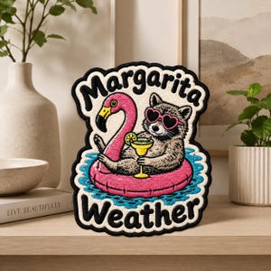 May include: An embroidered patch featuring a raccoon wearing heart-shaped sunglasses, relaxing in a pink flamingo pool float on blue water, holding a yellow margarita glass. The text Margarita Weather is stitched in black above and below the image.