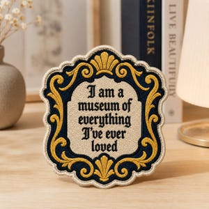 May include: A decorative embroidered patch featuring a cream-colored center with the black text 'I am a museum of everything I've ever loved' surrounded by a dark blue and gold ornate frame with a scalloped edge and a light beige border.
