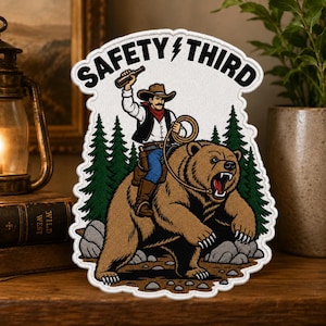 May include: A white embroidered patch featuring a cowboy riding a brown bear in a forest. The cowboy wears a hat, vest, and blue jeans, holding a bottle and a lasso. The text SAFETY THIRD is arched at the top with a lightning bolt in the center.