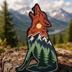 May include: An embroidered patch shaped like a howling wolf. The design features a layered landscape with a bright yellow sun in the orange upper section, white-capped dark blue mountains in the middle, and a green pine forest at the base.