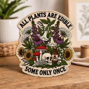 May include: An embroidered patch with a cream background featuring a botanical illustration of purple foxgloves, white flowers, sunflowers, and red mushrooms. A human skull and skeletal hand are depicted alongside a brown poison bottle.