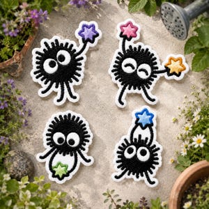 May include: Four embroidered patches featuring black, fuzzy, round creatures with large white eyes. One holds a purple star, one holds a pink and yellow star, one holds a green star, and one holds a blue star. Each patch has a white border.