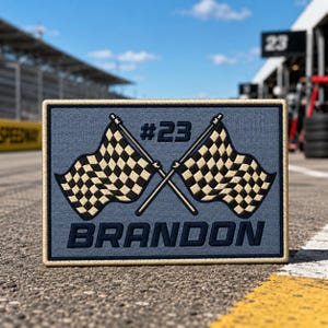 May include: A rectangular embroidered patch with a dusty blue background and a tan border. The design features two crossed black and white checkered racing flags, the text #23, and the name BRANDON in bold, dark blue capital letters.