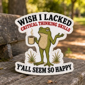 May include: An embroidered patch featuring a green frog holding a coffee mug with the text WISH I LACKED CRITICAL THINKING SKILLS and Y'ALL SEEM SO HAPPY. The frog has a tan belly and stands between two small patches of green grass.