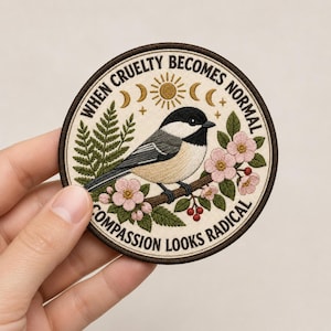 May include: A circular embroidered patch featuring a black-capped chickadee perched on a flowering branch with pink blossoms and green leaves. The text reads "WHEN CRUELTY BECOMES NORMAL COMPASSION LOOKS RADICAL" with a sun and moon design above.