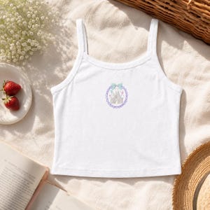 May include: A white sleeveless crop top featuring a central graphic print of a grey castle enclosed within a purple scalloped circular frame, topped with a light blue bow and small pink and blue balloons, finished with thin shoulder straps.
