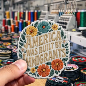 May include: Embroidered patch with the text "AMERICA WAS BUILT BY IMMIGRANTS" in tan thread. The patch is circular, framed by green leaves and colorful flowers in shades of yellow, orange, and teal. A stack of similar patches is visible.