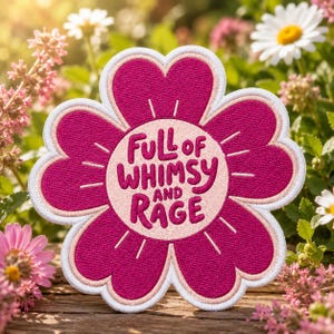 May include: A flower-shaped embroidered patch with dark pink petals and a light pink center. The text "FULL OF WHIMSY AND RAGE" is stitched in dark pink letters in the center, surrounded by small stitched lines radiating outward from the text.