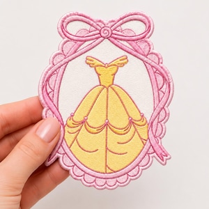 May include: An embroidered patch featuring a yellow ball gown with an off-the-shoulder neckline and a full skirt, centered within a pink oval frame decorated with a bow at the top. The design is stitched with vibrant yellow and pink thread.