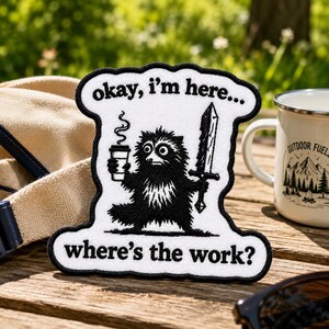 May include: A white embroidered patch with a black border featuring a black raccoon illustration holding a steaming coffee cup and a sword. The text reads, "okay, i'm here..." at the top and "where's the work?" at the bottom in black lettering.