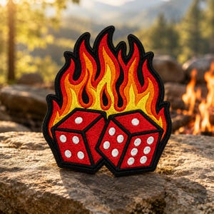 May include: An embroidered patch featuring two red dice with white pips positioned in front of a stylized flame design. The flames are colored in gradients of yellow, orange, and red, all outlined with a thick black border.
