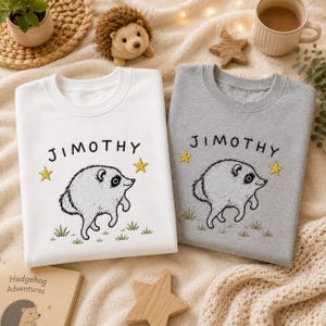 May include: A white sweatshirt and a gray sweatshirt, each featuring an embroidered illustration of a hedgehog, the name "JIMOTHY" in black text, two yellow stars, and green grass blades at the bottom of the design on the front chest.