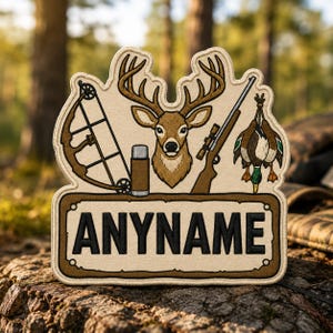 May include: A beige embroidered patch featuring a central deer head, a compound bow, a rifle with a scope, a thermos, and a hanging duck. Below these items is a rectangular banner with the black text ANYNAME centered in a bold font.